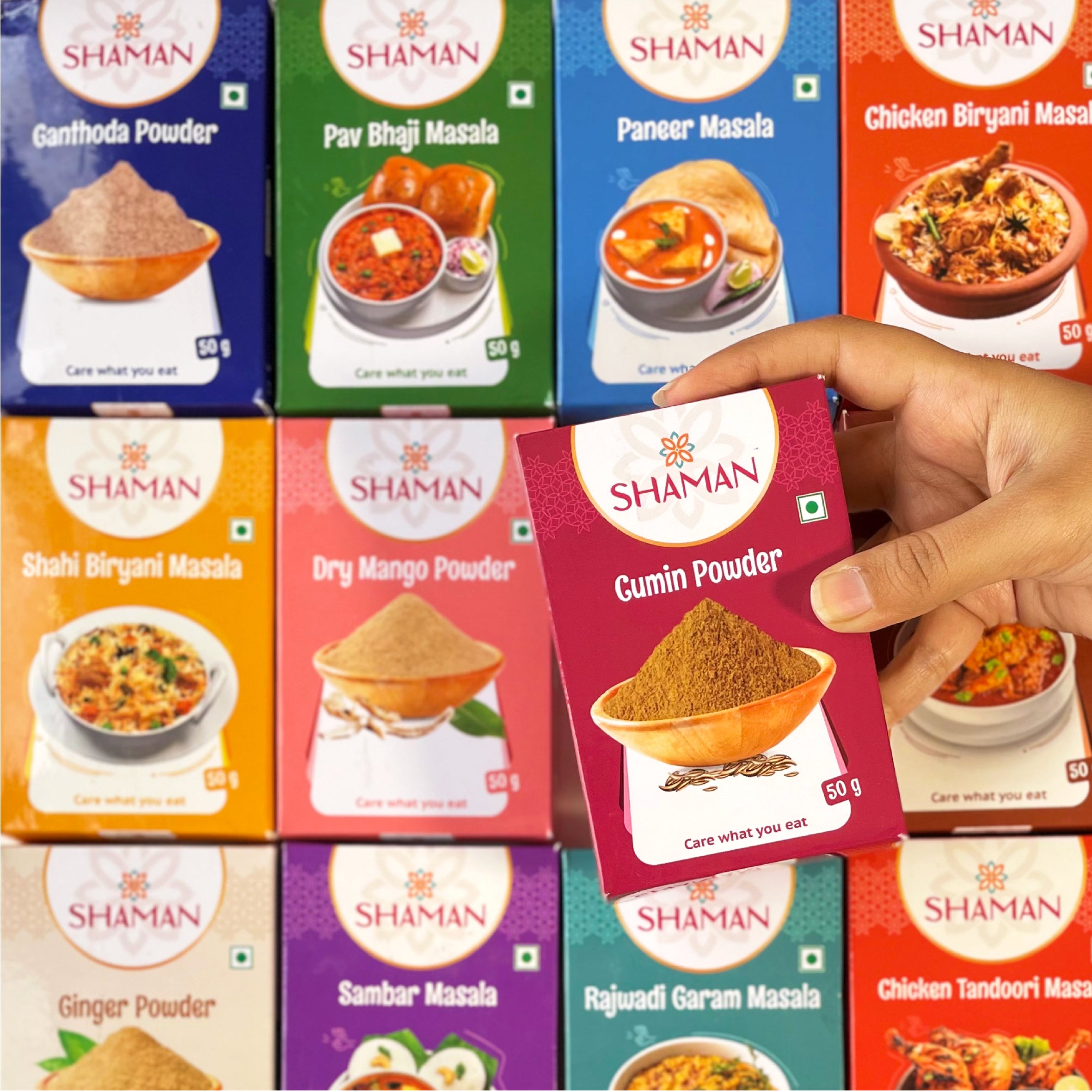 Hand holding a 'Shaman' cumin powder packet with various other 'Shaman' spice packets in the background.