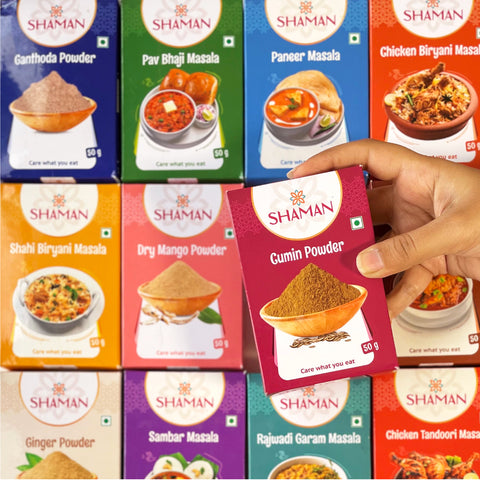 Hand holding a 'Shaman' cumin powder packet with various other 'Shaman' spice packets in the background.